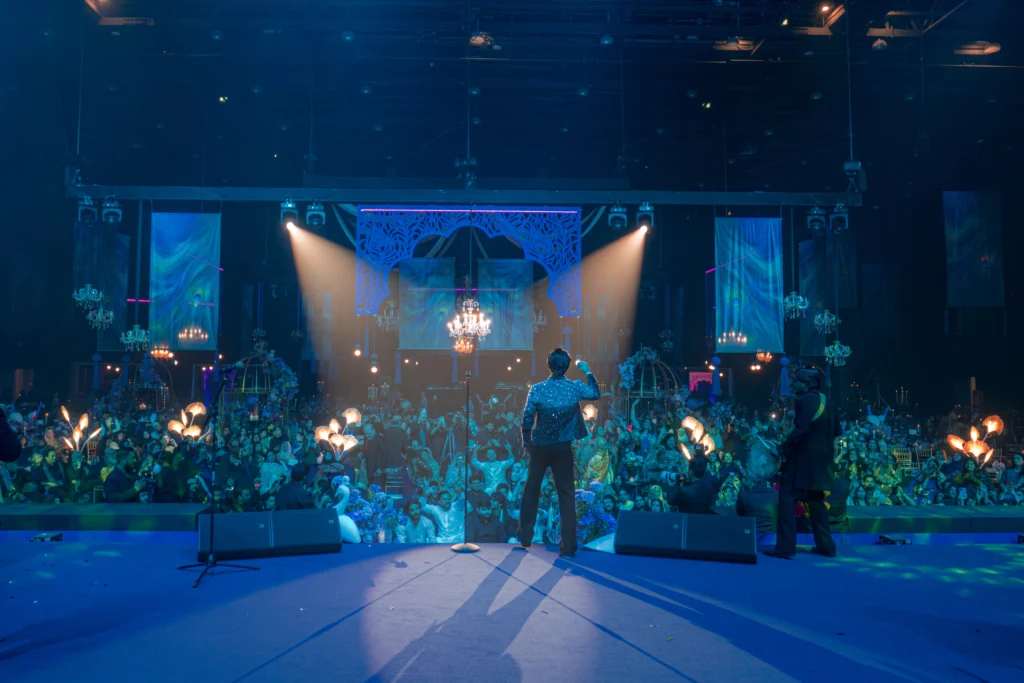A live performer facing a packed wedding crowd at Madinat Jumeirah with an ornate arch backdrop, crystal chandeliers, decorative floral torch lights and vibrant blue stage lighting