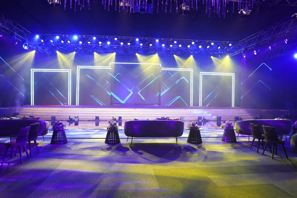 A stunning Megawatt Live event stage with blue and yellow LED lighting, geometric neon backdrop and stylish lounge seating arrangement