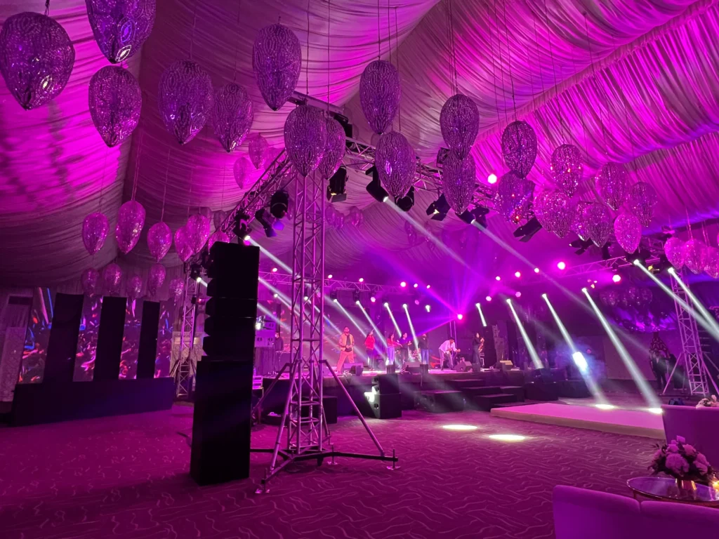 A luxurious outdoor wedding marquee with pink and purple LED lighting, decorative hanging lanterns and a live performance on stage