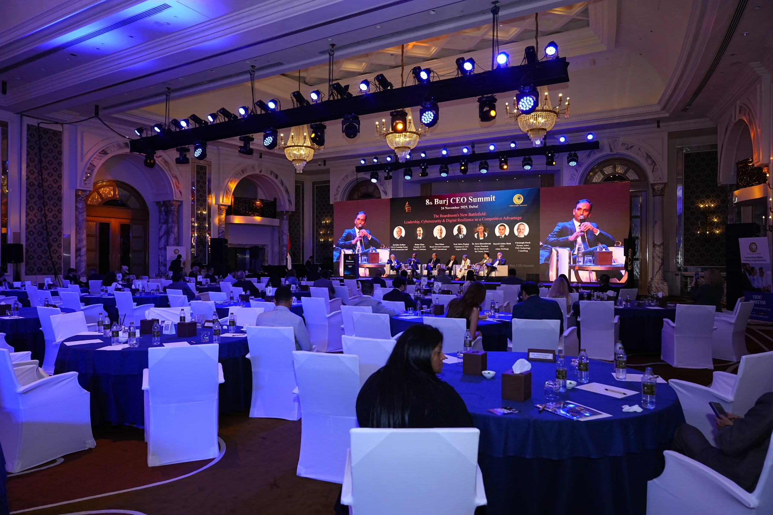 8th Burj CEO Summit panel discussion at a luxury Dubai ballroom with a large branded LED screen, crystal chandeliers and professionally arranged conference dining tables