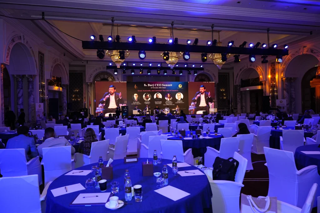 8th Burj CEO Summit panel session at a grand Dubai ballroom with a large branded LED screen, crystal chandeliers, blue ambient lighting and neatly arranged conference dining tables