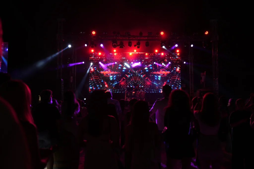 A energetic outdoor concert at Barasti Beach with a packed crowd, vibrant red and blue LED stage lighting and a stunning geometric video wall backdrop