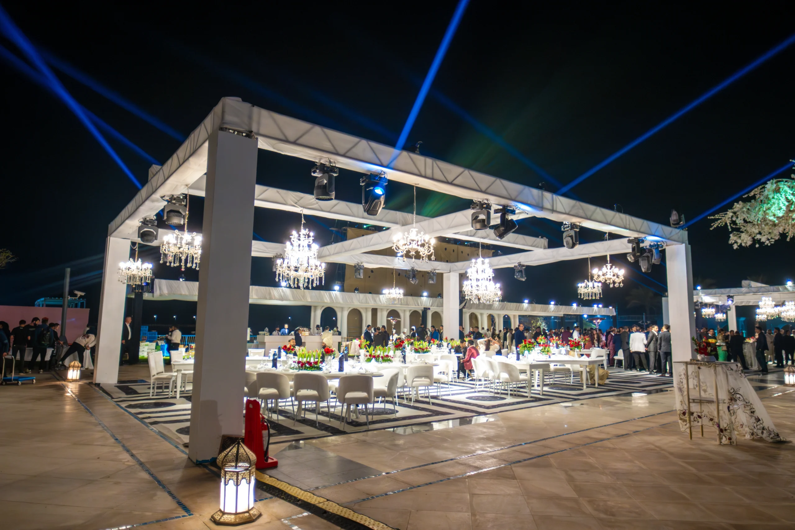 An elegant outdoor Qatar wedding dinner under a white pergola with multiple crystal chandeliers, floral table centerpieces and stunning blue laser lights in the night sky