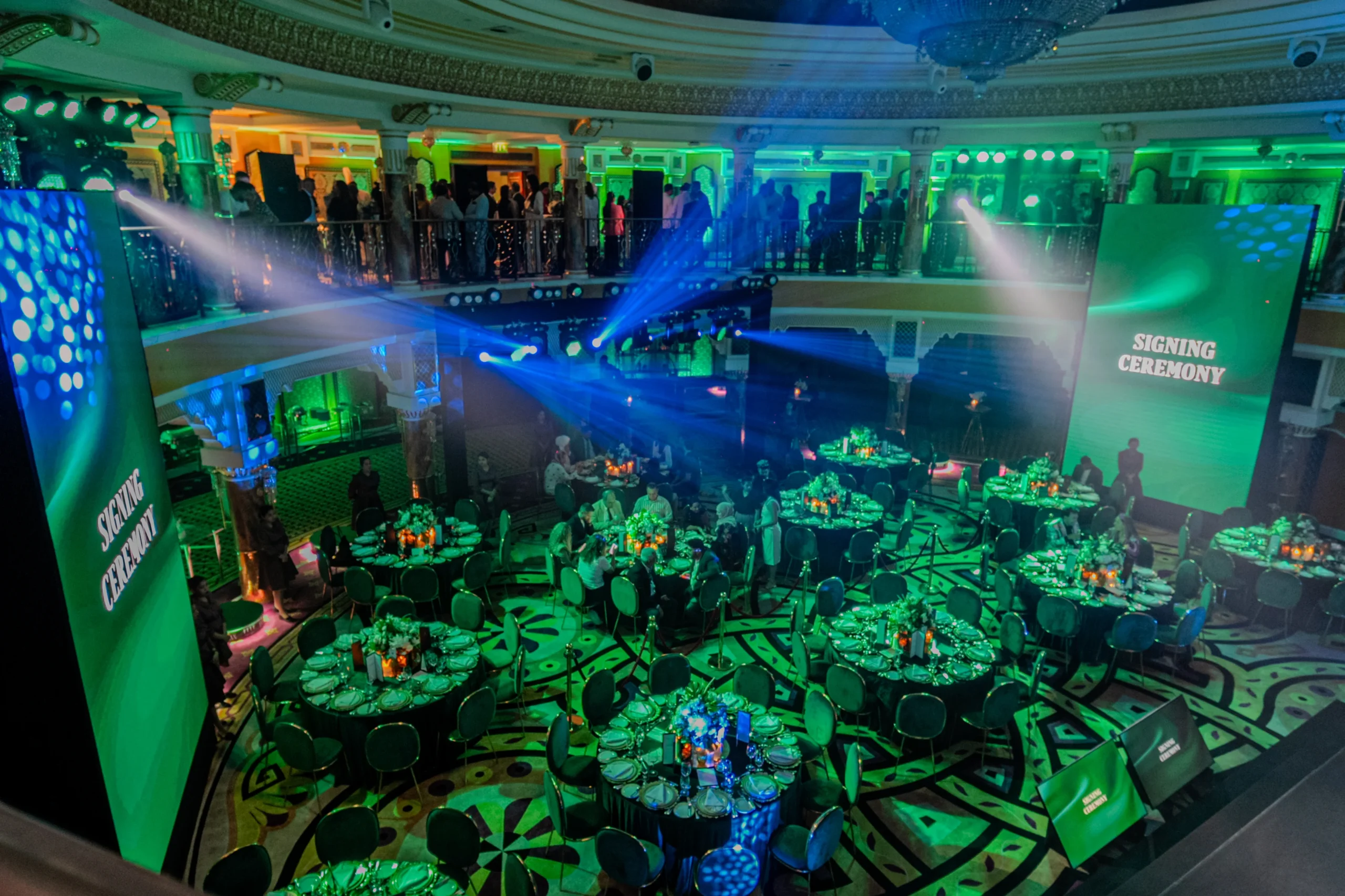 A formal signing ceremony at a luxury Dubai venue with vibrant green lighting, LED screens, round dining tables and guests on a stunning double-level balcony